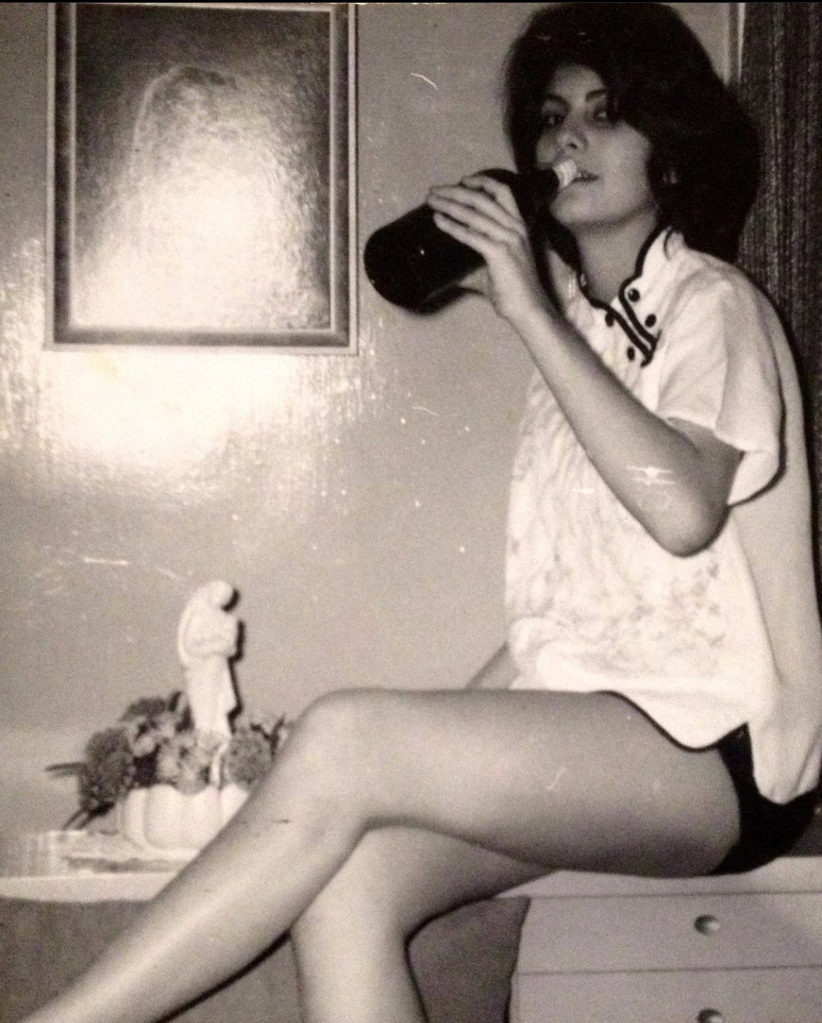 an old black and white photograph of a women (Judith) sitting on a dresser drinking a bottle of wine with her legs crossed